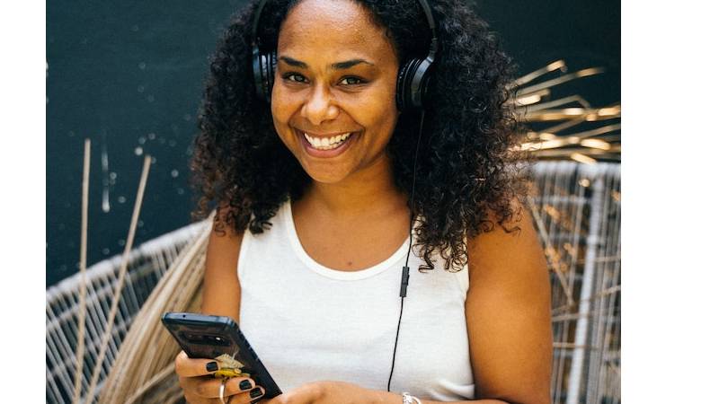 A smiling person sits on a chair while wearing headphones and holding a mobile phone