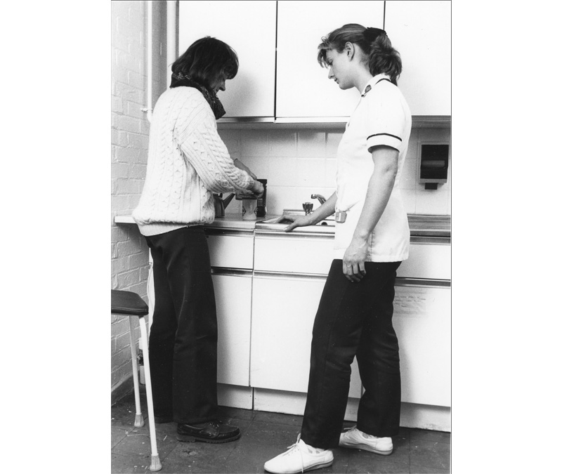 Black and white photograph of an occupational therapy student standing with a patient while she makes a hot drink.