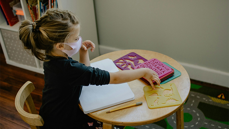 Image of a young girl sitting at a table wearing a face covering, playing with stencils. 