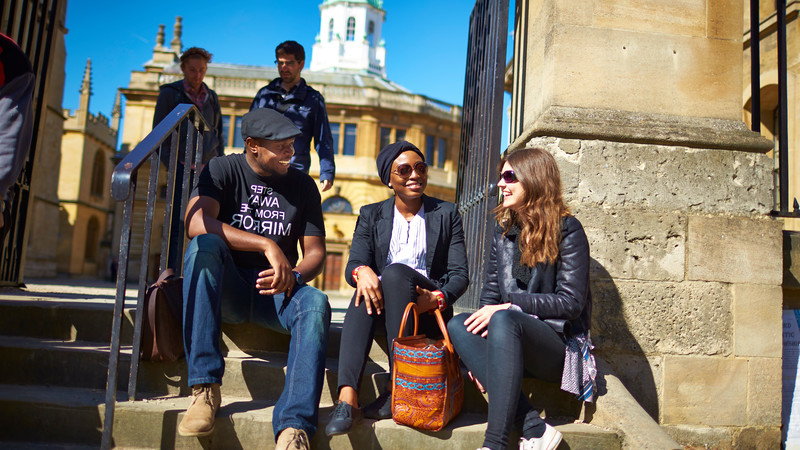A group of people sitting on the sandstone steps of an Oxford landmark.