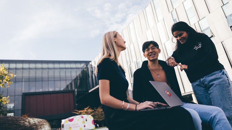 Three students outside on campus with a laptop having a conversation