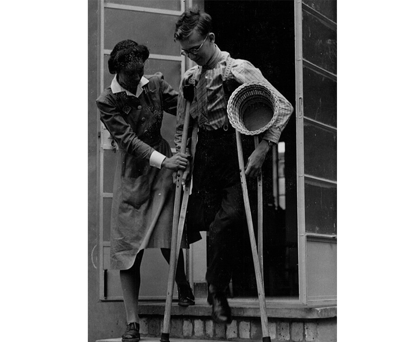 Black and white photograph of an occupational therapy student helping a man on crutches to walk down a step.